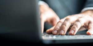 Low-angle view from the back of the computer of a businessman typing on a laptop computer keyboard.