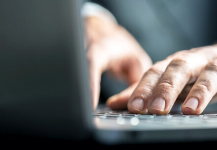 Low-angle view from the back of the computer of a businessman typing on a laptop computer keyboard.