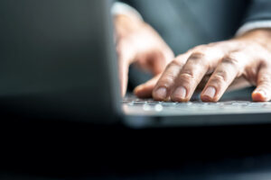 Low-angle view from the back of the computer of a businessman typing on a laptop computer keyboard.