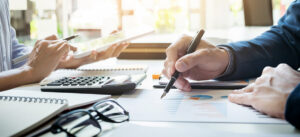 two businesspeople sit across a table from each other working on financial reports.